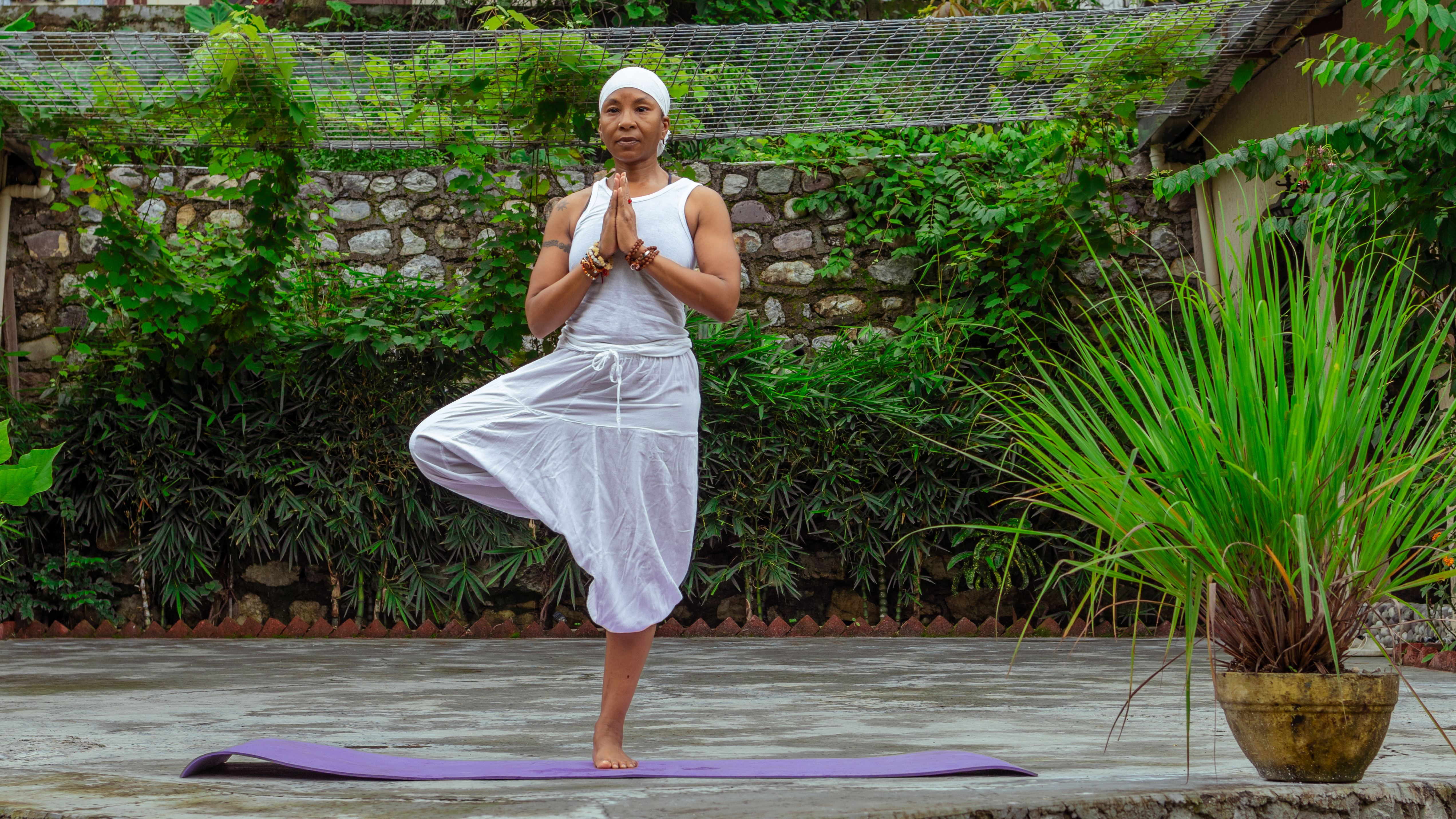 A woman in white flowing trousers and a vest top practices the one-legged yoga tree pose on a mat surrounded by greenery.