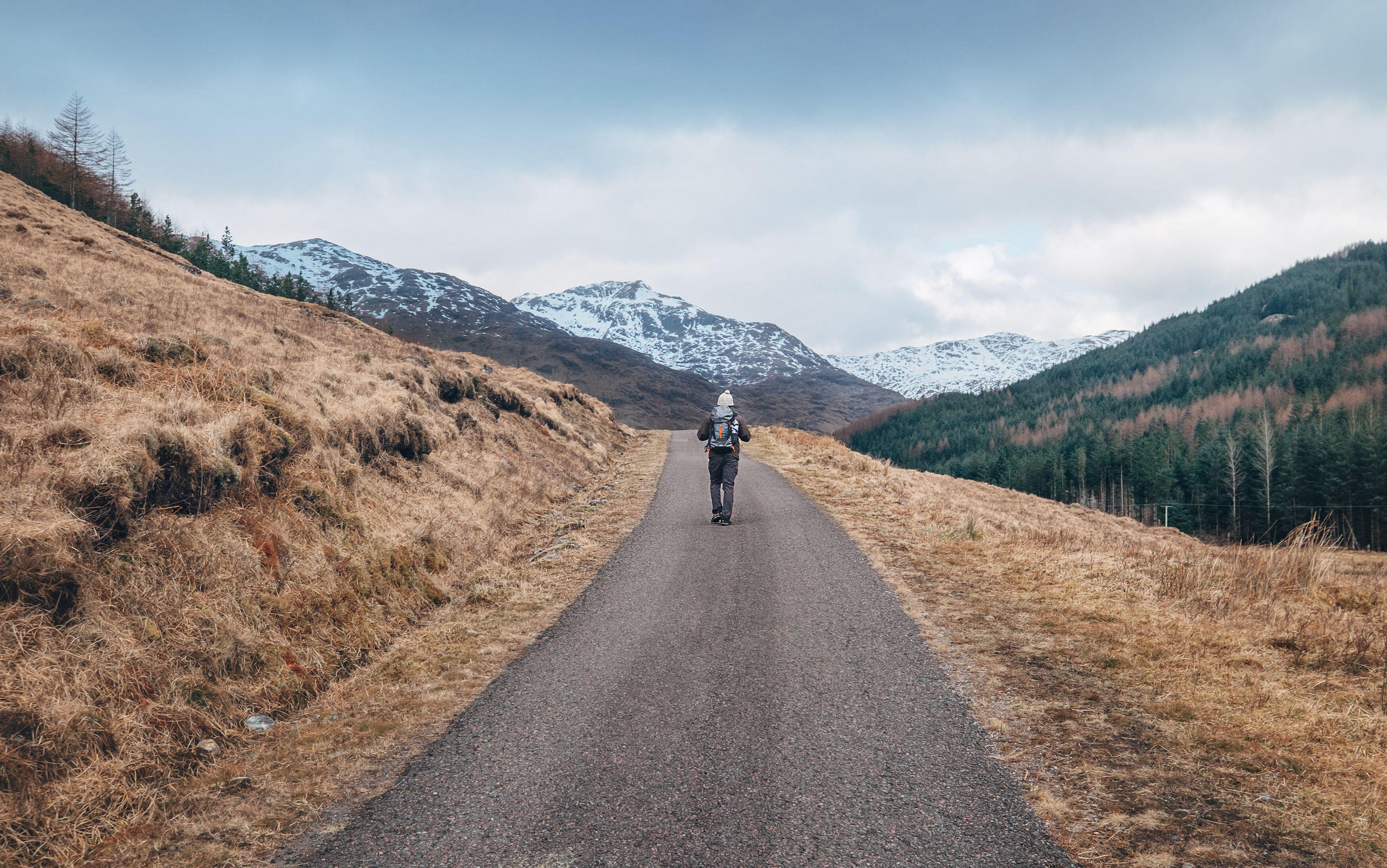  Lonely backpacker man walking with backpack by Scottish Highlands mountain road with snowy peaks at early moody morning.