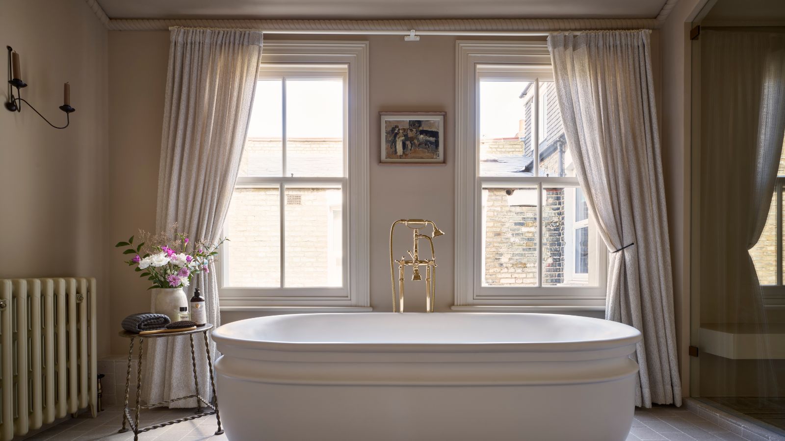 A warm neutral bathroom with a freestanding bath and floor length curtains over the windows