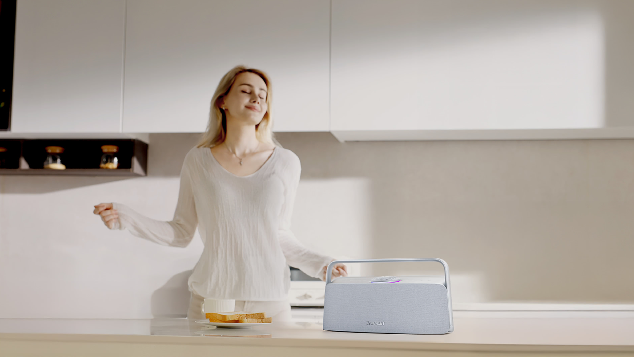A young woman dancing in her kitchen with a Tronsmart Fiitune X30 speaker on the worktop in front of her.