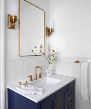 A white bathroom with a gold mirror, gold sconces, a white and blue sink, and a gold towel rail