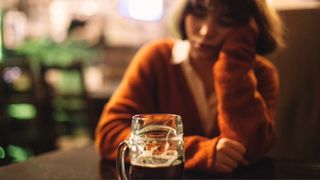 A woman in rust orange jumper falling asleep with head in hand sat behind a pint