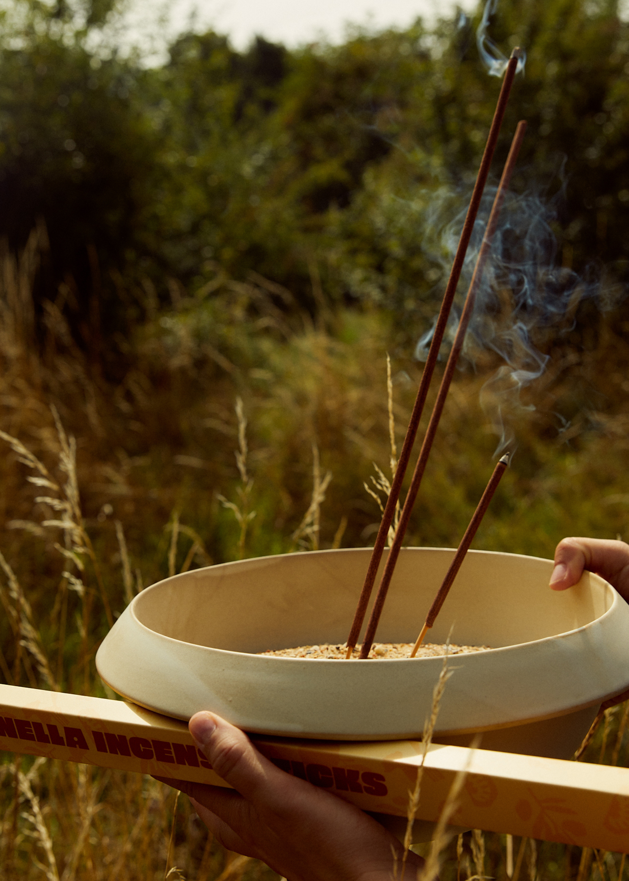 A pair of hands holding an incense bowl as it smokes in a field