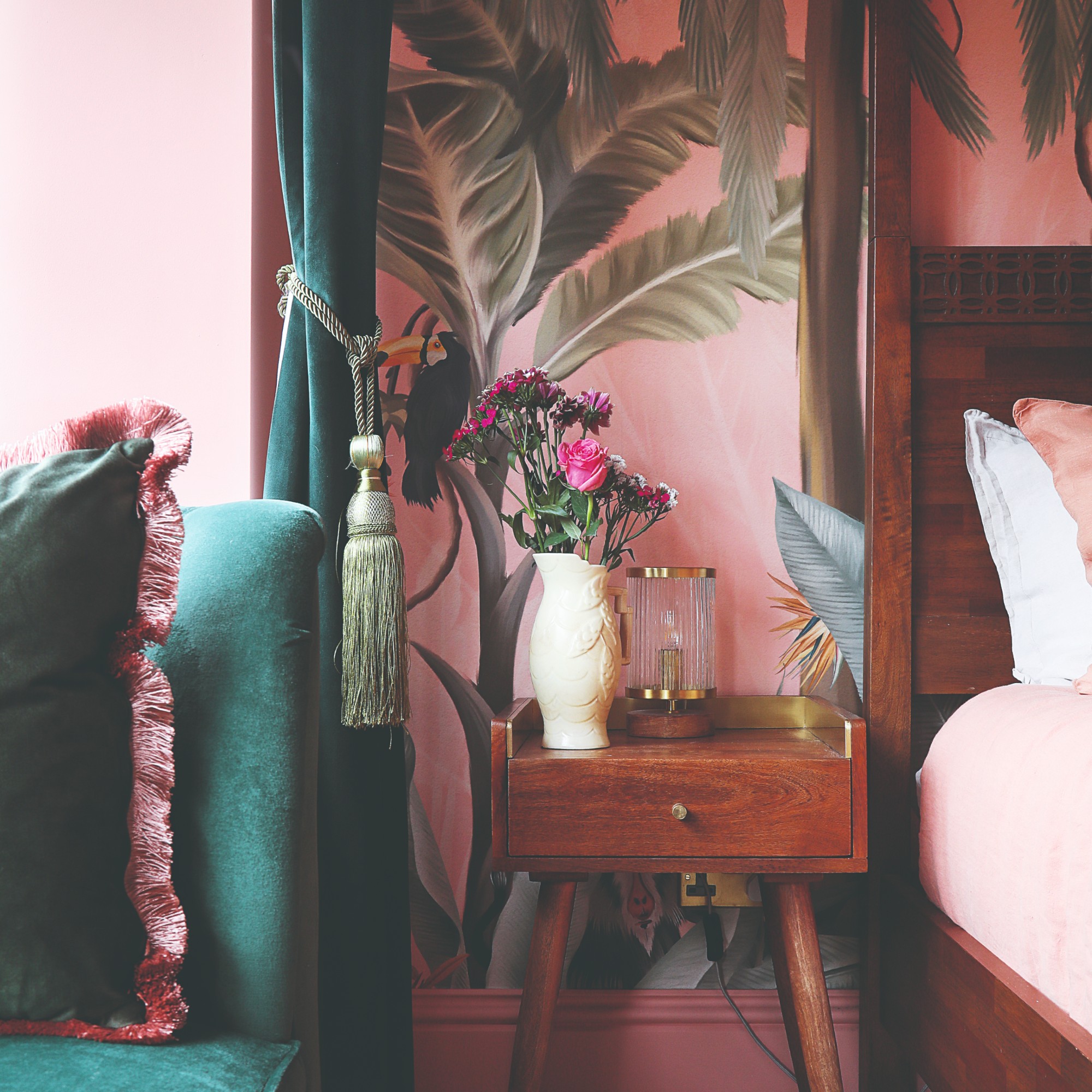 A bedroom with a pink palm leaf-print wallpaper, a tassel curtain tie on a dark green velvet curtain and a fringe-trimmed cushion
