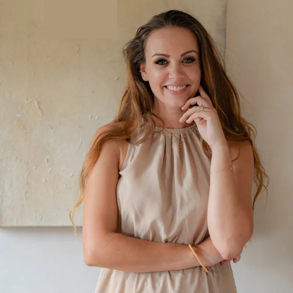 female with long brown hair wearing beige dress stood against neutral wall colour