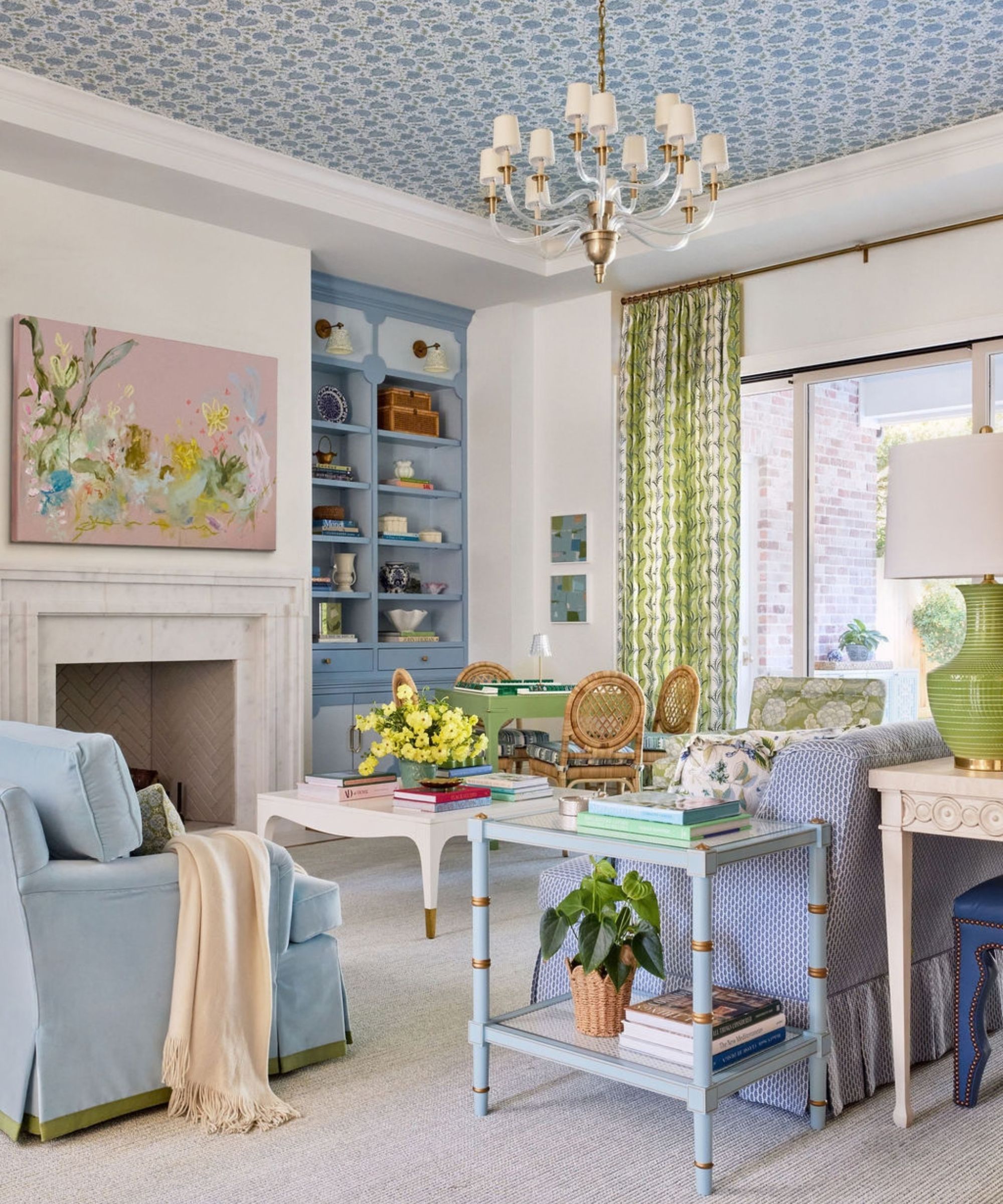 Airy living room featuring a fireplace, blue and green seating, a blue wallpapered ceiling with a central chandelier, and built-in blue shelving