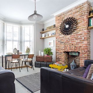 Living room with an exposed brick wall fireplace with a star shaped clock at the top of the wall, open wood shelves, light walls and carpet with dark furniture and a yellow foldable coffee table