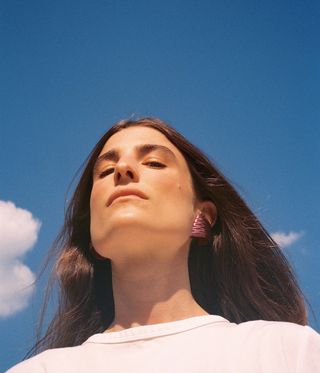 Maria Sole Ferragamo modelling dark pink earrings, hair flowing, white tshirt, blue sky and white soft clouds backdrop