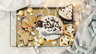 The the best (and worst) festive foods for sleep image shows a golden tray of gingerbread cookies, mince pies and marshmallow-topped hot chocolates placed on a bed dressed with faux fur blankets