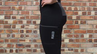 Side view of rider standing in front of a wall wearing Pedaled Odyssey Women's Cargo Bib Tights