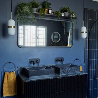 A dark blue ensuite bathroom with navy wall paint, soft rectangular mirror, vanity with double sink setup and circular metal towel rails holding mustard yellow towels. A shelf overhead displays indoor houseplants in grey planters with candles