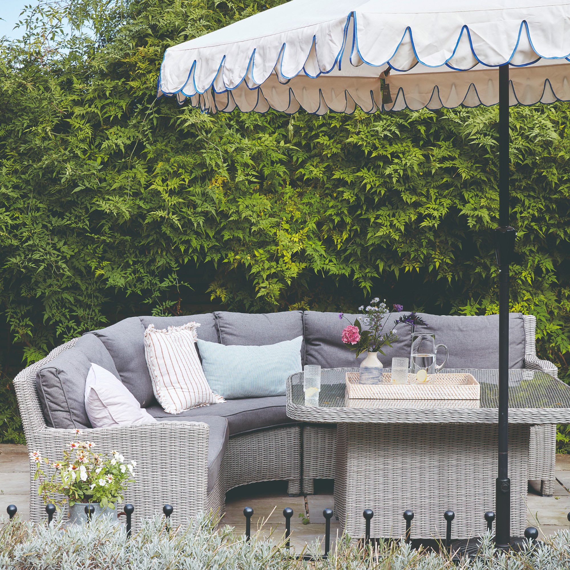 A garden with a grey rattan corner outdoor sofa and a matching table with a scalloped parasol next to it