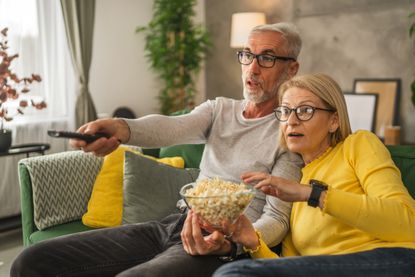 Mature senior couple husband and wife watch scary movie and hold popcorn with remote control at home
