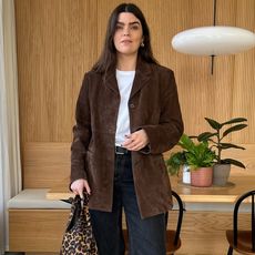 British style influencer Anna Newton poses in her chic London dining room wearing a chic outfit with a brown suede jacket, white T-shirt, black belt, black jeans, and a leopard-print bag