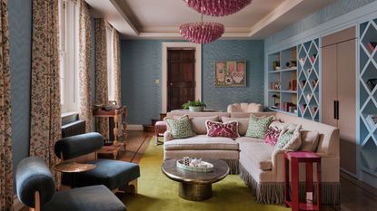 a blue wallpapered home library with a long wall of bookcases, a corner settee with fringing, pink chandeliers and side table