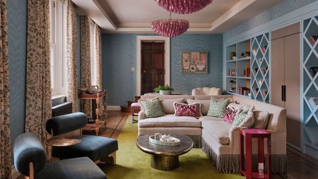 a blue wallpapered home library with a long wall of bookcases, a corner settee with fringing, pink chandeliers and side table