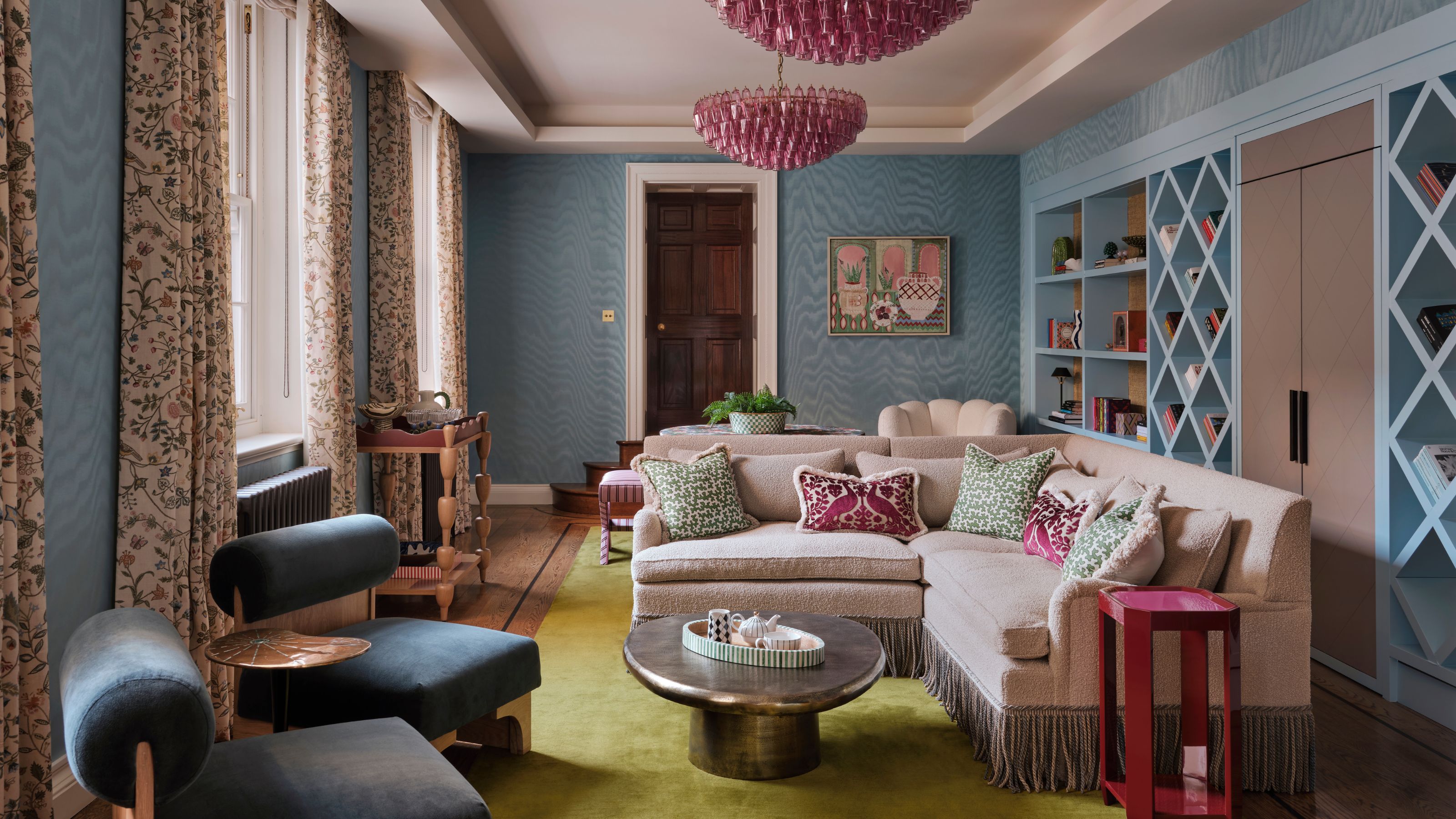 a blue wallpapered home library with a long wall of bookcases, a corner settee with fringing, pink chandeliers and side table
