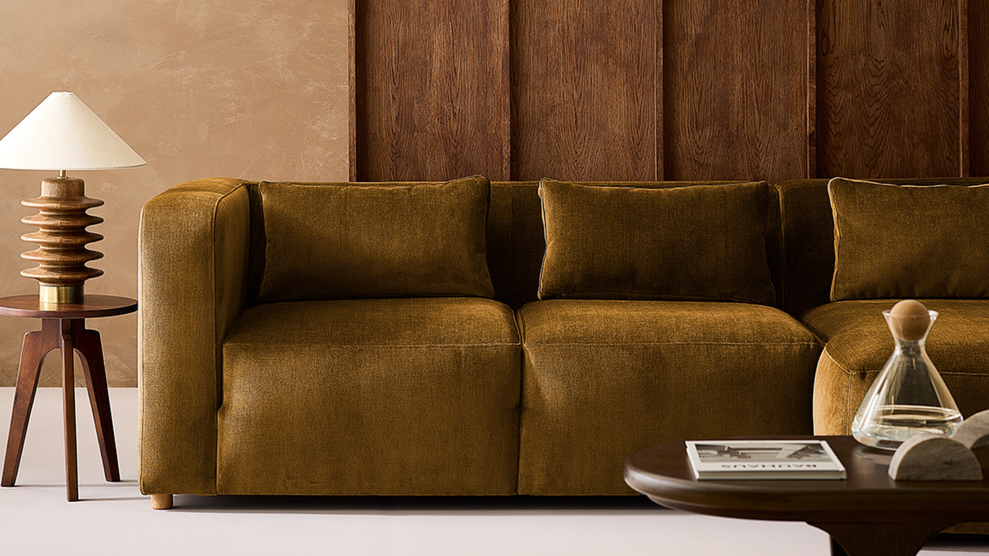 Image of a living room with beige walls and dark wooden paneling. There is an olive green, velvet sofa and a small wooden side table with a wooden accent lamp on it. 