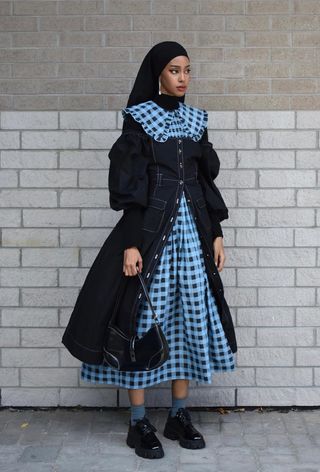 A woman's jean dress outfit with a puff-sleeve denim midi styled over a gingham-print dress with black loafers and blue socks.