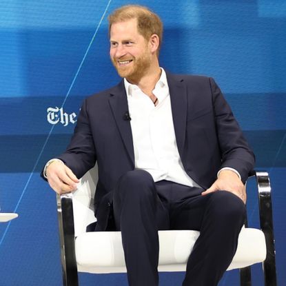 Prince Harry wearing a blue suit and white shirt sitting in a white chair in front of a blue New York Times background