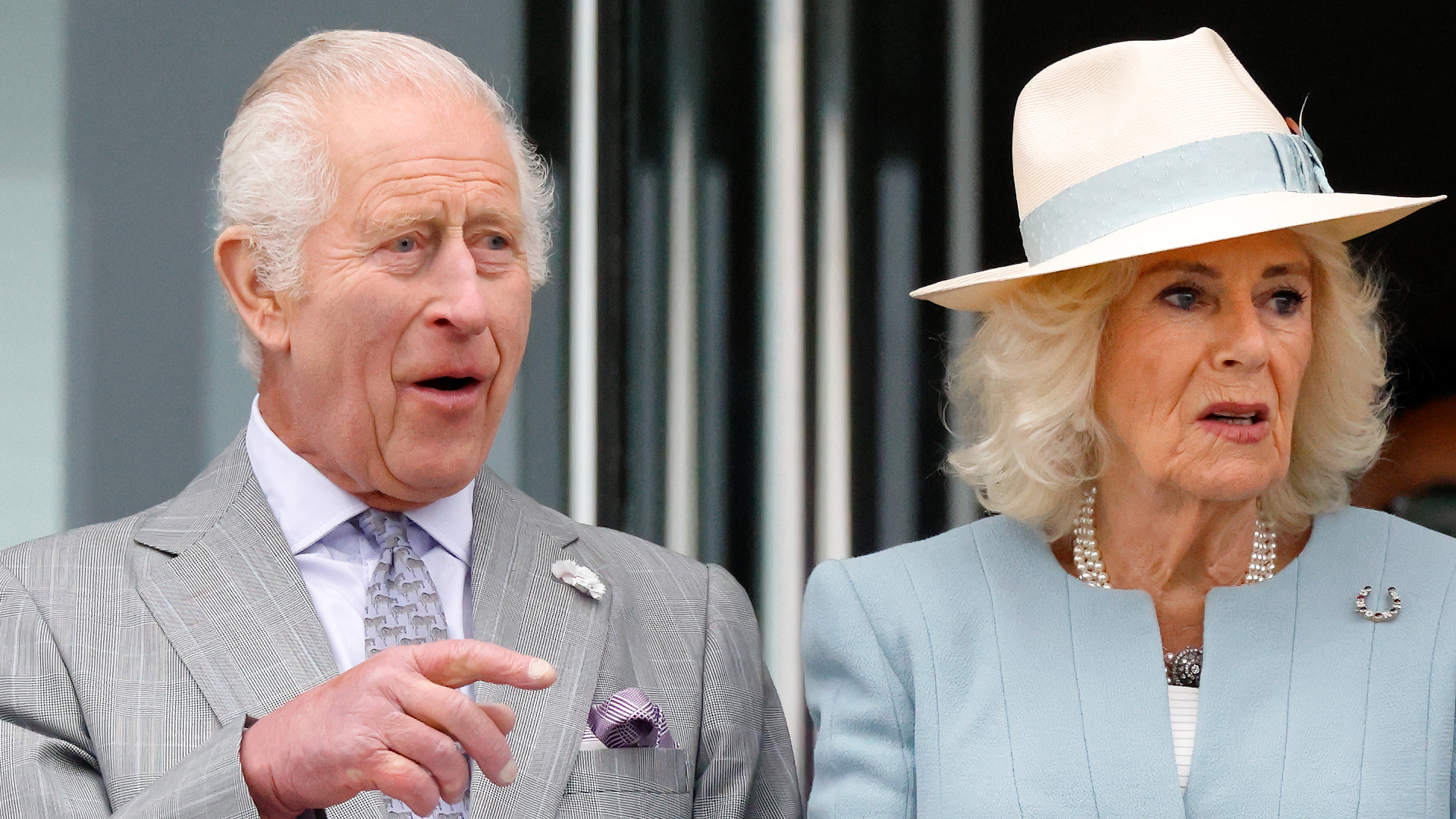 King Charles wears a gray suit and looks shocked while wife Queen Camilla wears a light blue suit jacket