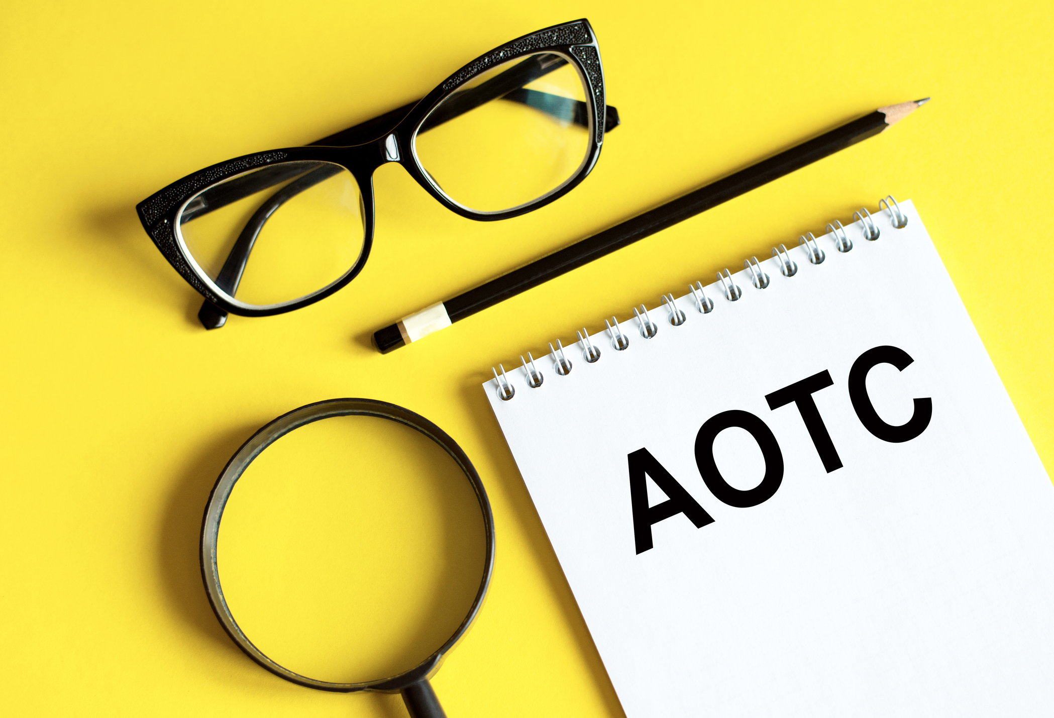 &quot;AOTC&quot; written on a notepad with glasses, a pencil and a magnifying glass on a yellow background