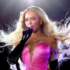 Beyonce wears a pink strapless dress with a low-cut V-neckline, with long black gloves, and her blonde wavy hair blows in the air