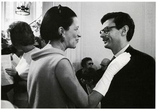 A woman having a conversation with a man at a party with her hands dressed in white gloves on his chest
