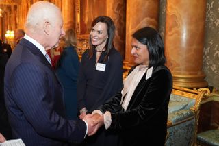 King Charles wearing a blue pinstriped suit shaking hands with Naveed Chowdhary-Flatt who is wearing a black velvet blazer and cream pussybow blouse