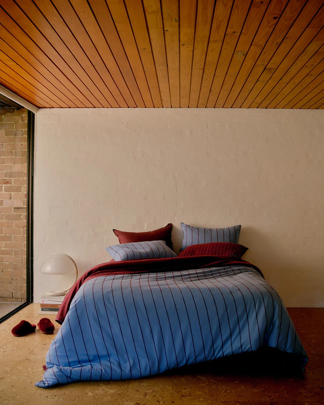 A bed in a minimalist room with white walls and cork floors. The bedding is blue and maroon stripes. 