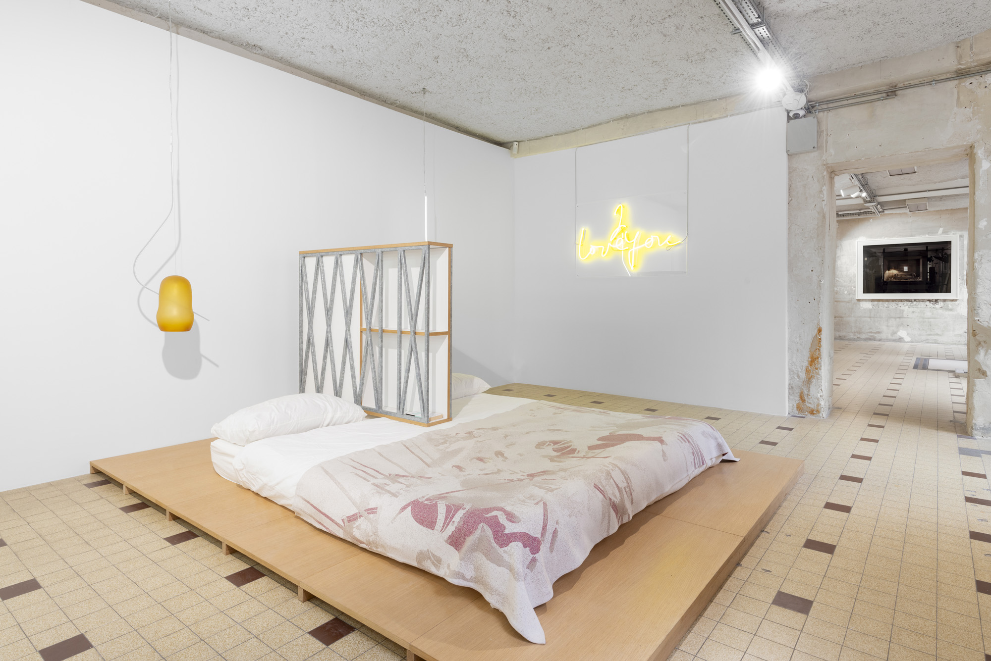 A bedroom made of a futon-style mattress with a wooden base, pink-shaded sheets, and neon lights is recreated inside an industrial gallery space with bright lighting and checkered tiles.