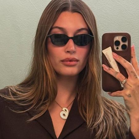 Hailey Bieber wearing sunglasses, a brown blazer, and her B necklace.