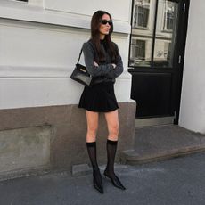 Woman wears a black pleated skirt, grey cardigan, sheer knee socks and pointed black slingbacks. 