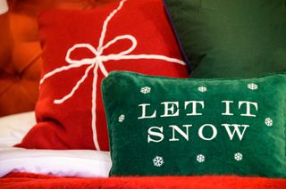 A green pillow that says 'Let It Snow' and a red cushion that has a white ribbon embroidered on to look like a present.