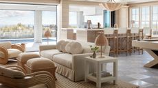 An open-concept living space where a tailored linen sofa and rattan lounge chairs meet a custom light-oak pool table, all set against a backdrop of dunes and sea.