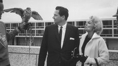 A black-and-white photo from 1960 of Barron and Marilyn Hilton looking at a large bird and its handler in front of a hotel.
