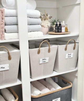 Organized linen cupboard interior in chic neutrals, with labelled baskets of different linen categories.
