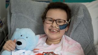 photo of a young smiling girl wearing black-framed glasses and holding a blue teddy bear while sitting up in a hospital bed 