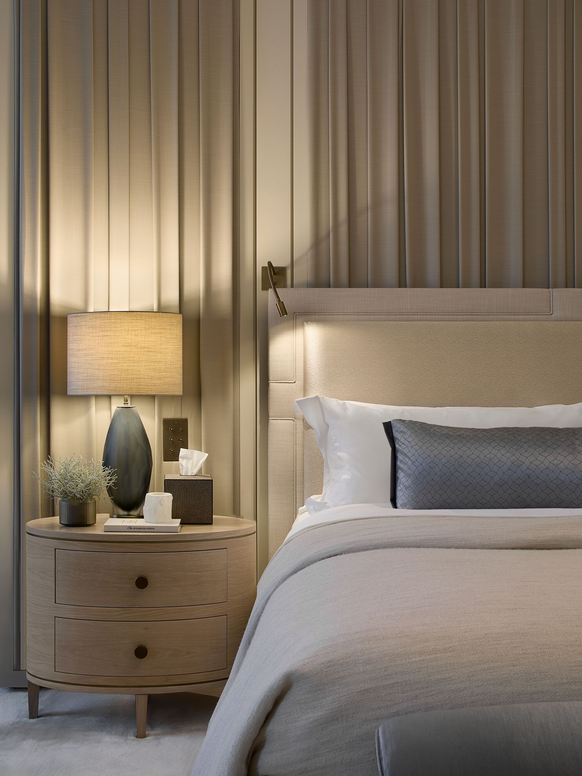 modern bedroom with curtains behind the bed, upholstered bedframe with wall sconce, round side table with lamp, candle, box of tissues, and potted plant, with ottoman at the end of the bed
