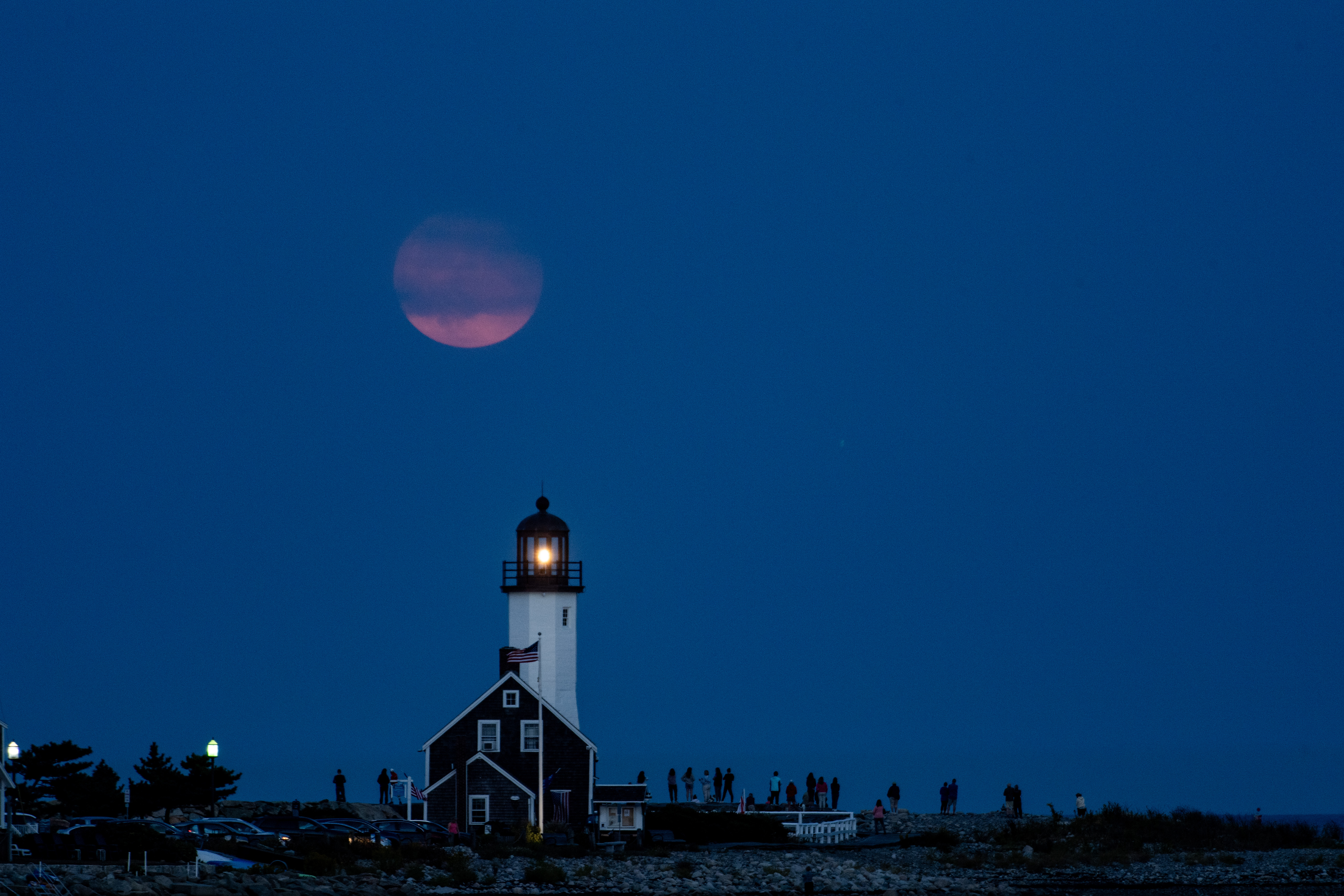 dusky pink colored moon rising in blue sky over a small lighthouse painted white with a red top.