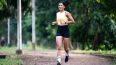 A woman runs along a dirt path with trees lining either side. She wears a vest, biker shorts, a sports watch, sneakers and long socks.