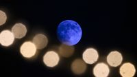 A blue moon is pictured in a black sky with an out of focus string of lights arcing below in the foreground.