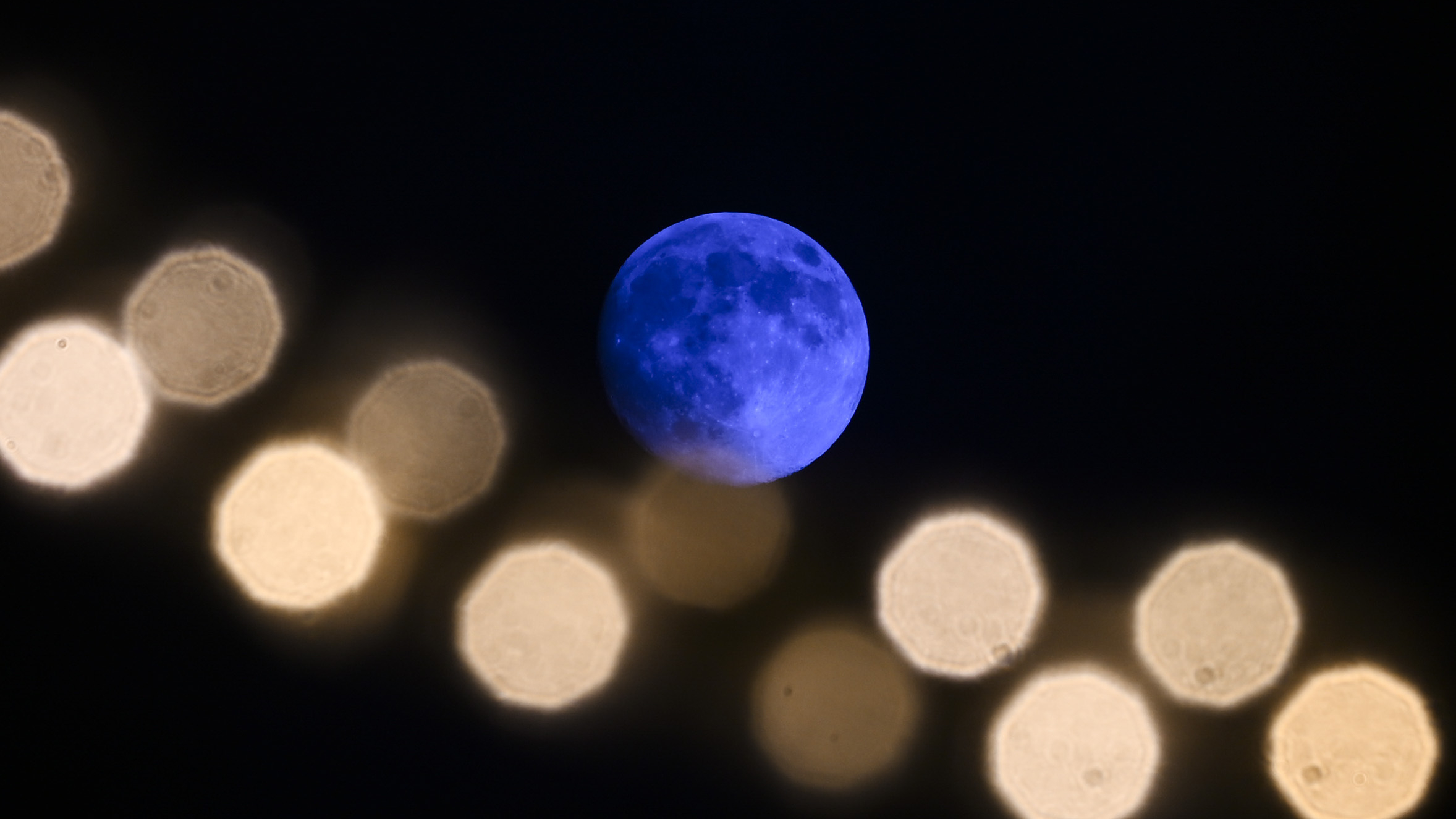 A blue moon is pictured in a black sky with an out of focus string of lights arcing below in the foreground.