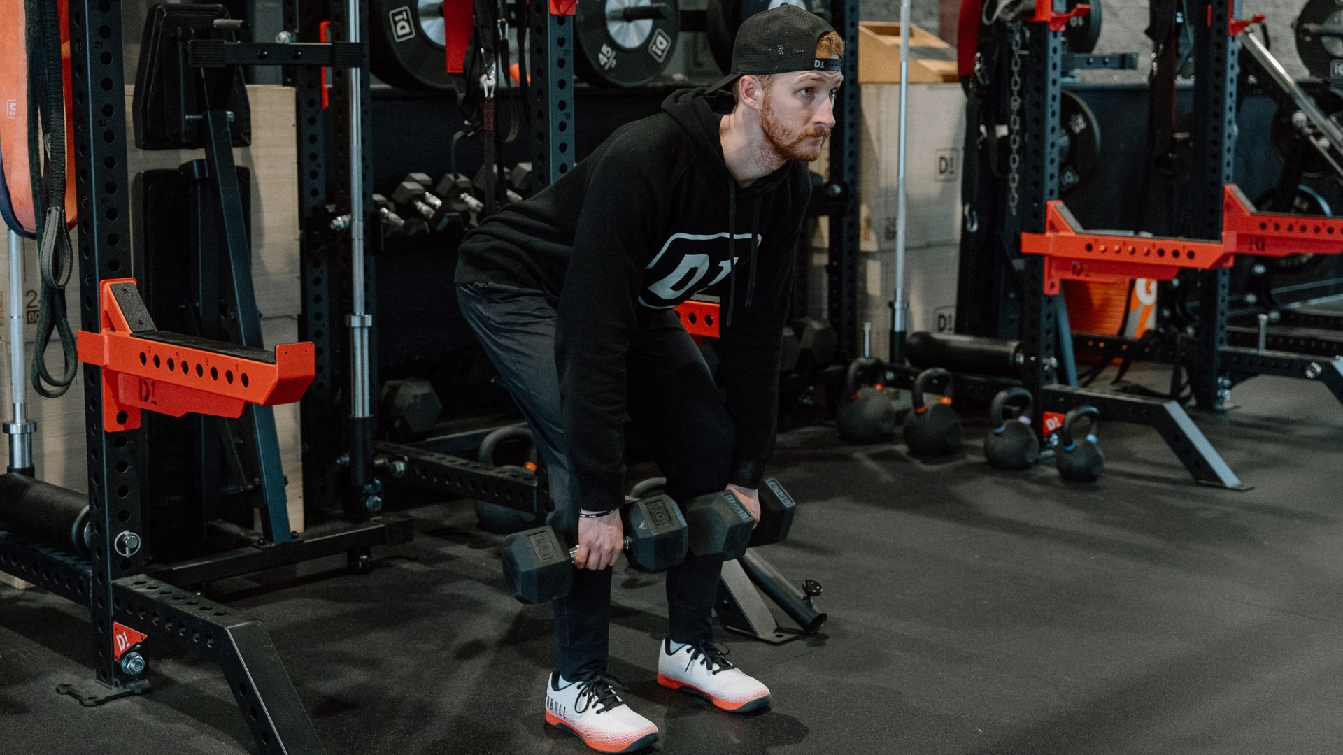 man in black hoodie and trousers performing a Romanian deadlift with two dumbbells and a squat rack behind him.