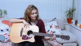 Woman sat on a couch, fretting the third fret of her Fender acoustic guitar