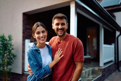 A couple hugs and poses in front of their home, or in the backyard.