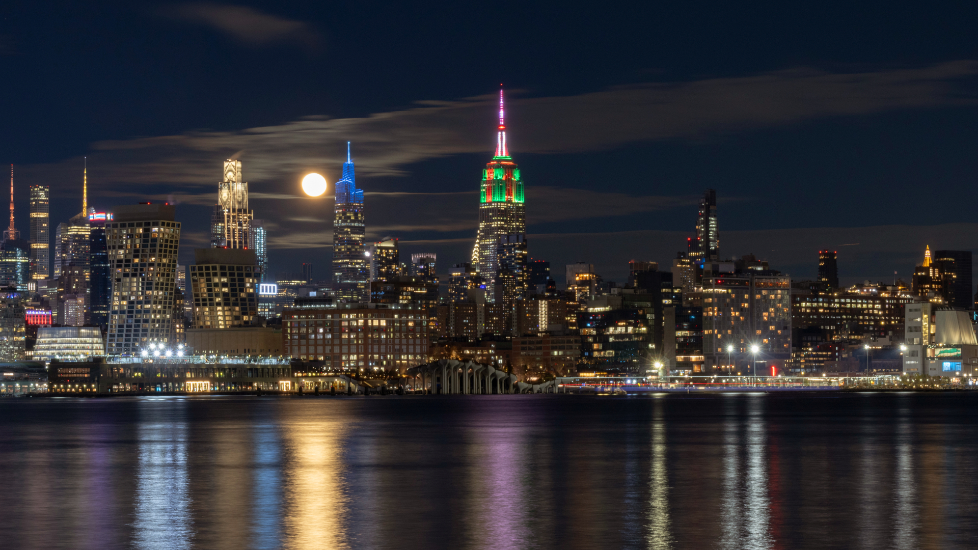 The yellow glowing disk of the moon is pictured shining between skyscrapers on the New York skyline at night, as light from the natural satellite and the city casts columns of illumination across a foreground body of water.