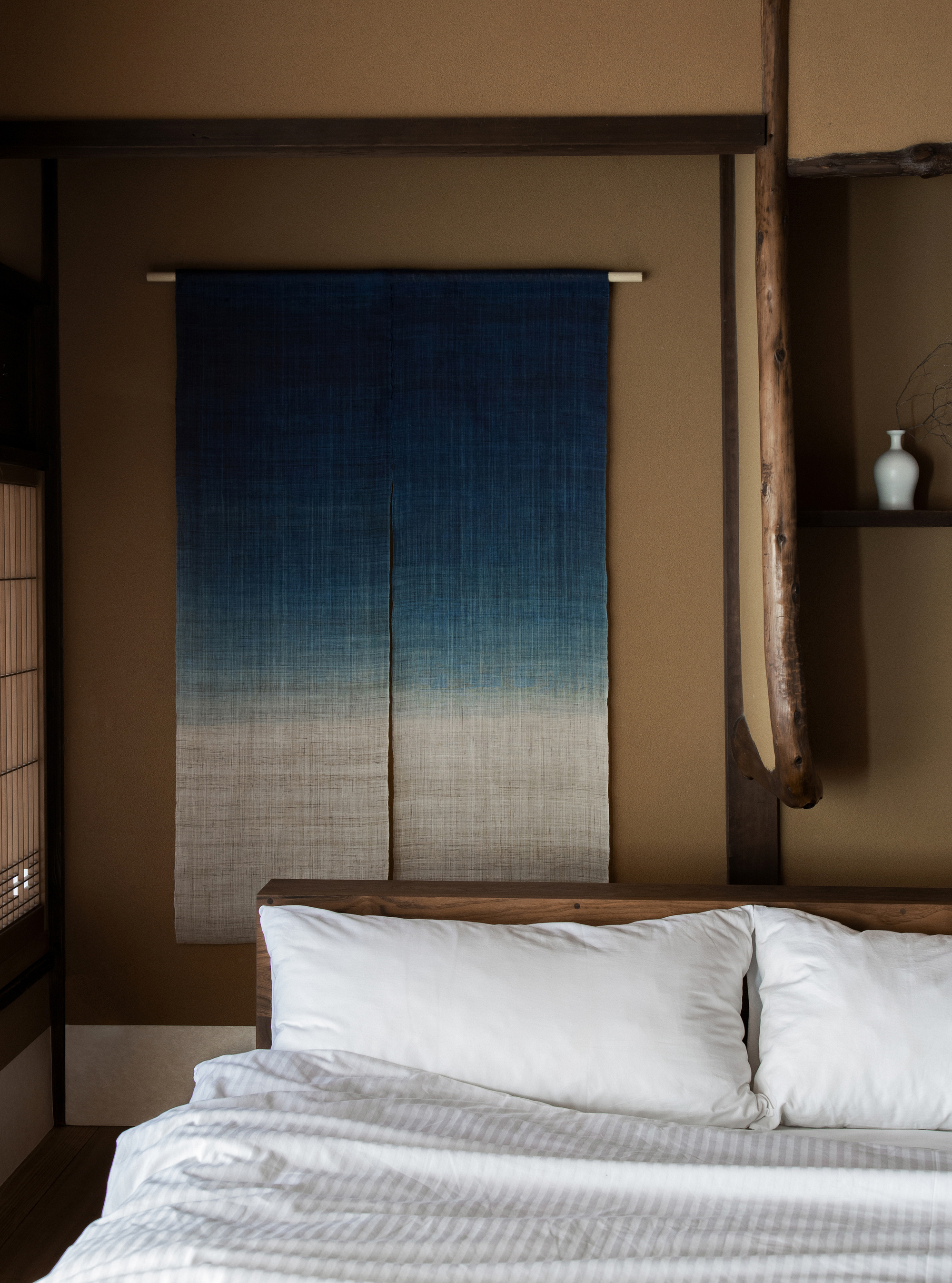 Image of a blue and white gradient noren hanging on a dark tan wall in a minimal bedroom that has a bed with white bedding.