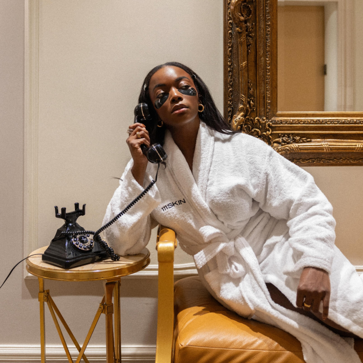 A woman wears skincare eye patches and talks on the phone.