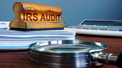 A wooden IRS Audit stamp sits atop a stack of papers next to a calculator and magnifying glass.